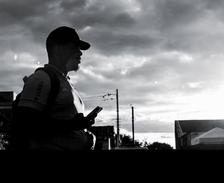 Samuel Moraes de perfil, com boné e celular na mão, silhueta em preto e branco sob céu dramático